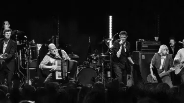 Black and white photo of a lively folk band performing on stage. Musicians play diverse instruments, including guitar, accordion, and banjo.
